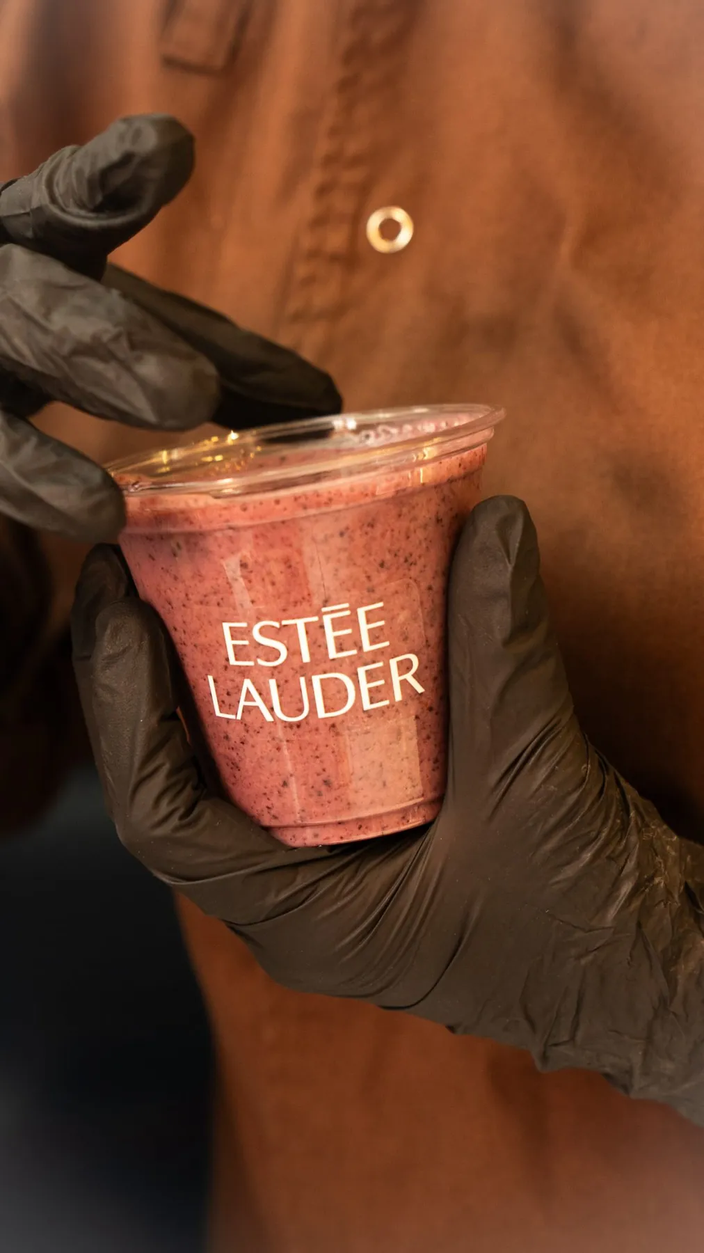 Barista con guantes negros sostiene un vaso con el logo de Estée Lauder en una activación de marca.