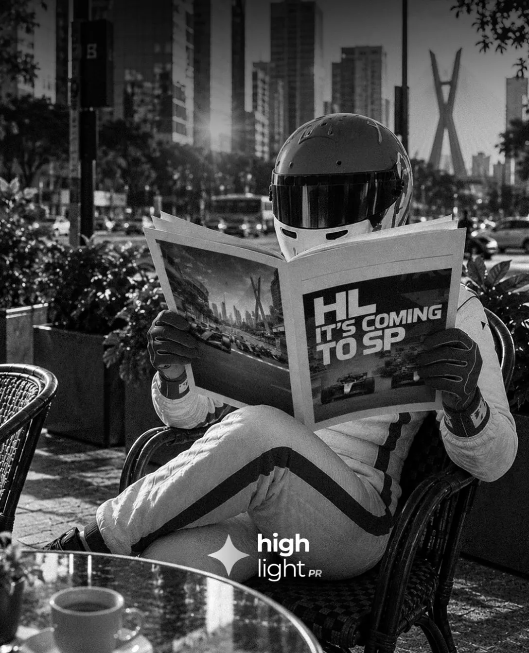 Piloto con casco lee un diario con el titular «HL it's coming to SP» en una terraza de São Paulo, en blanco y negro.